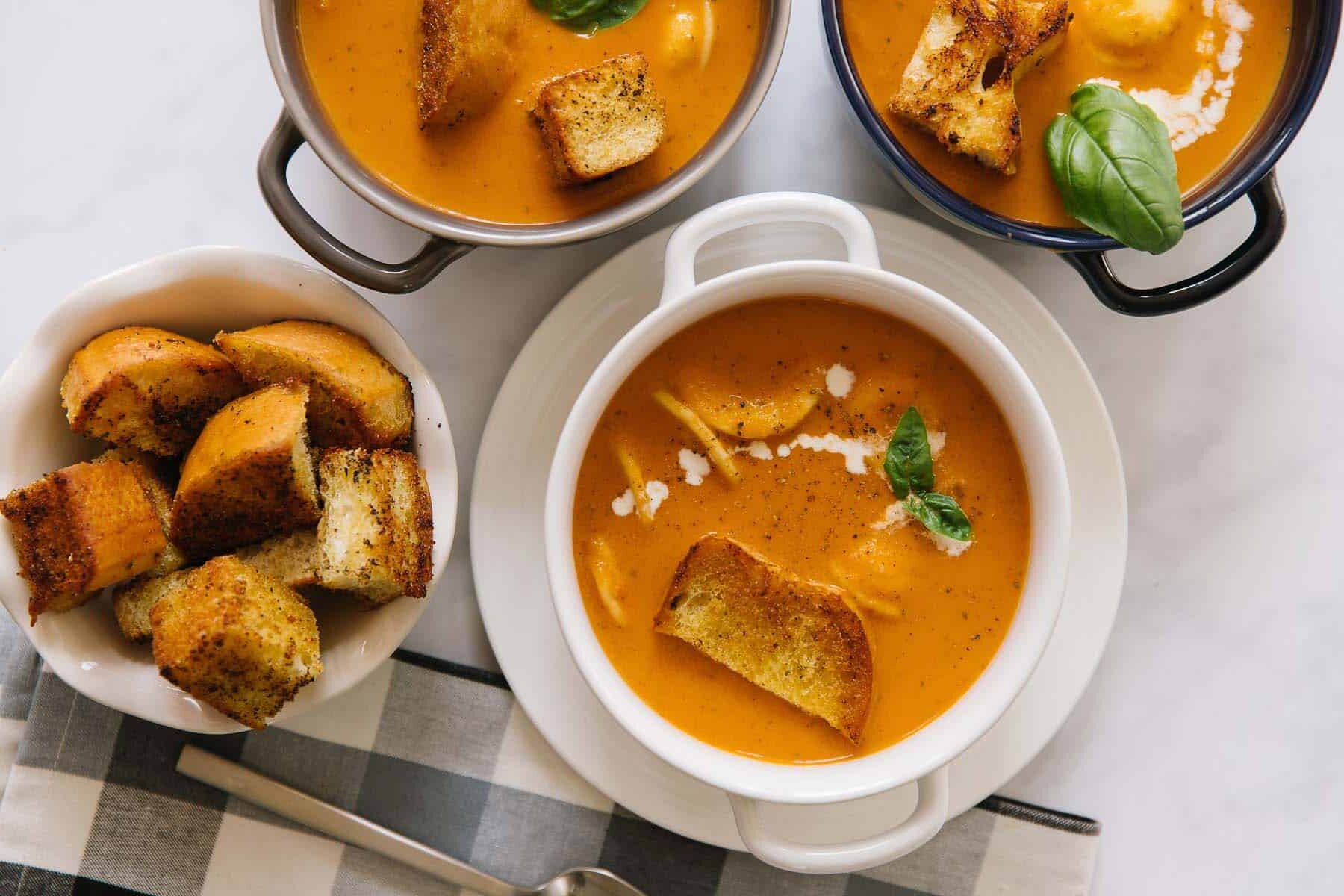 A white, black and silver pot of tomato basil soup topped with croutons and a cream drizzle with homemade croutons on the side.
