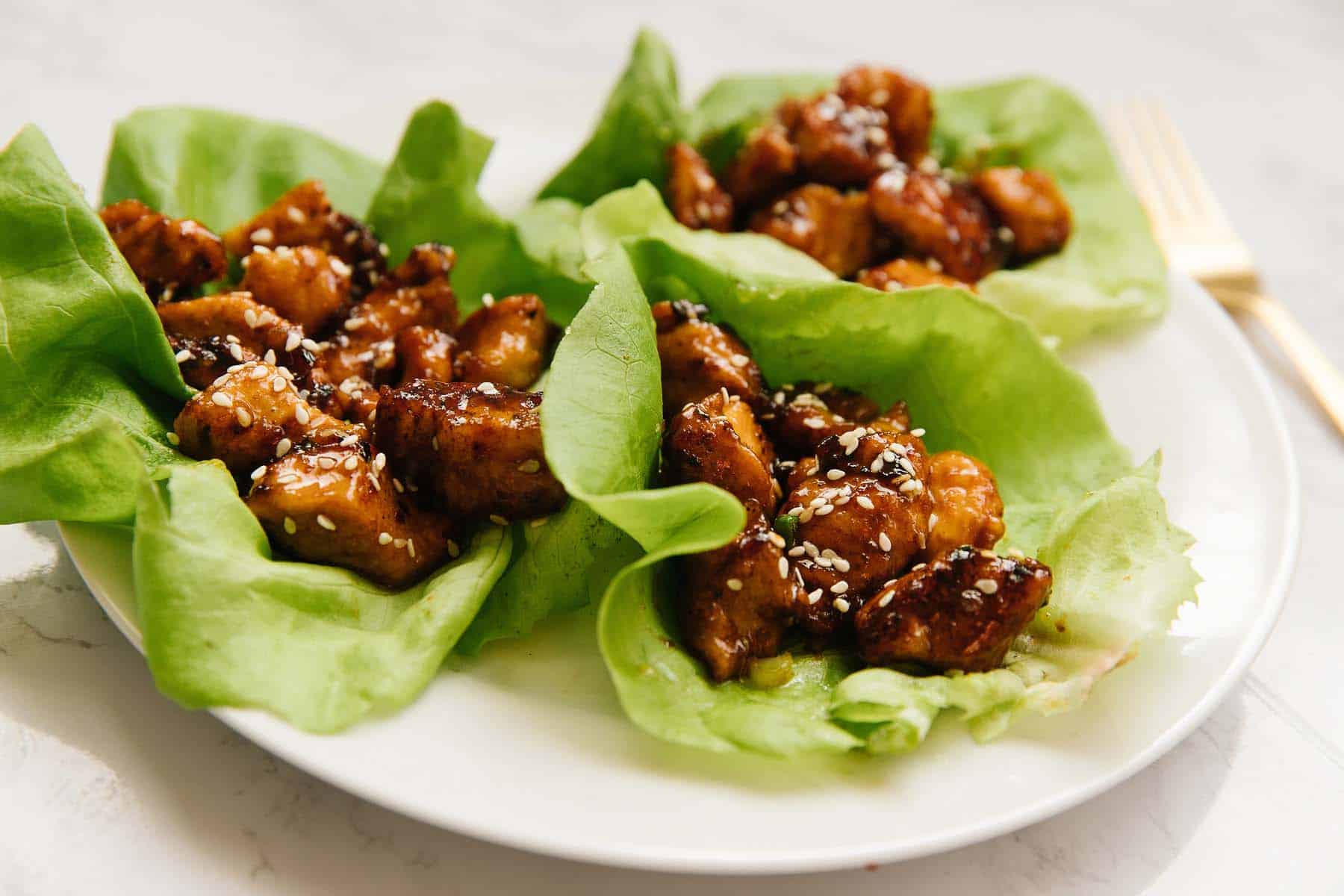 A white plate with three lettuce wraps filled with sesame honey chicken mixture with a gold fork on the side.