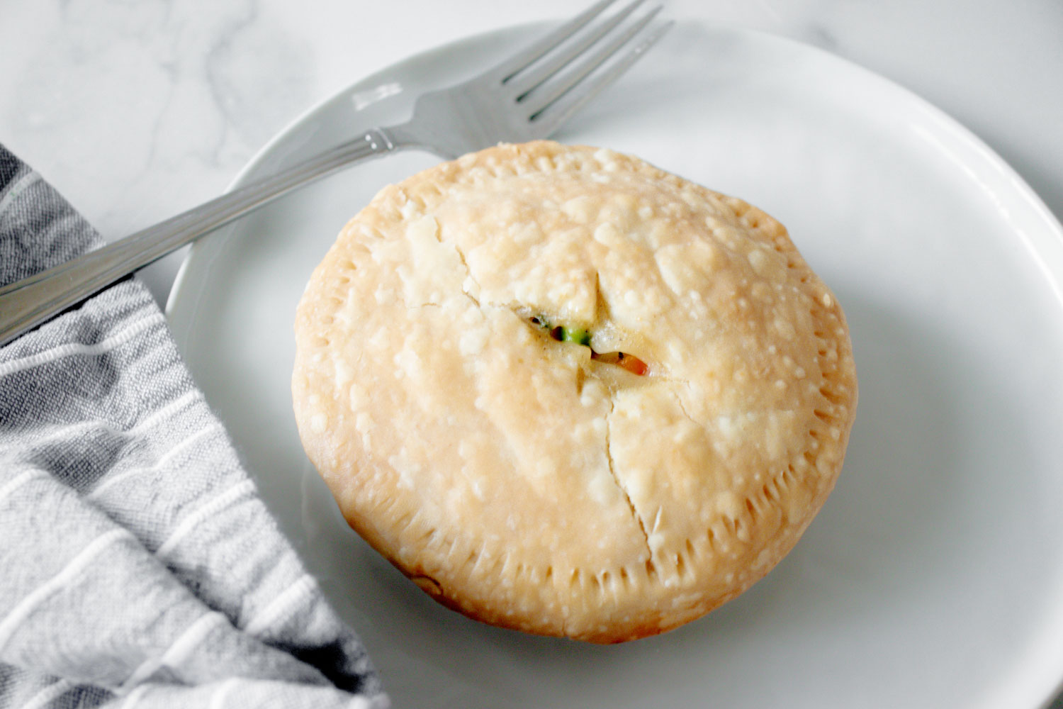 One mini chicken pot pie on a white plate with a silver fork and a gray and white striped towel on the side.