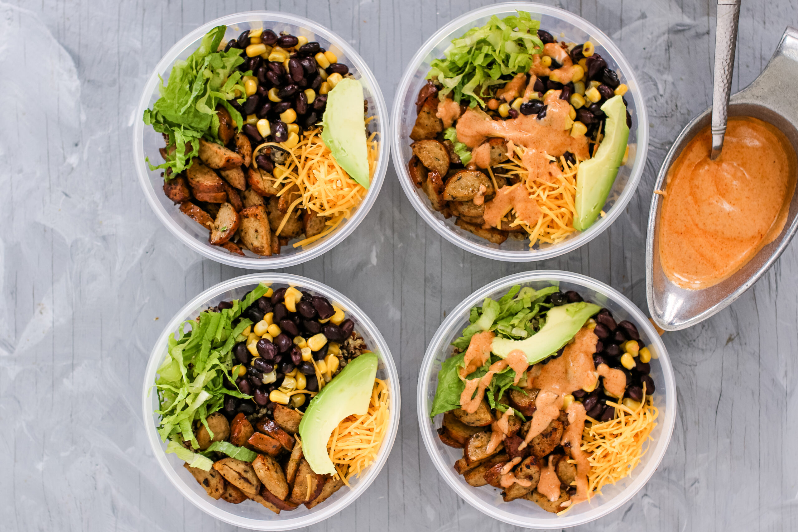 Four plastic bowls of quinoa burrito bowls and toppings with a silver bowl of sauce on the side.