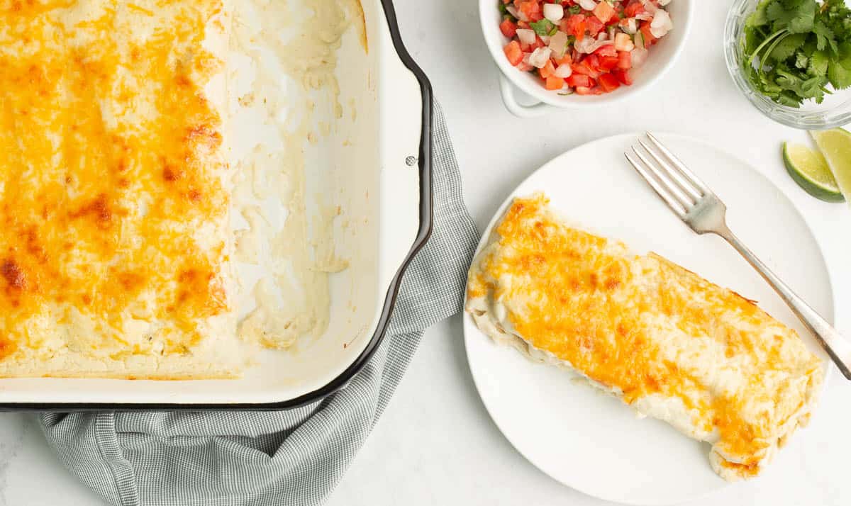 A white plate with one enchilada with a silver fork on it with a pan of enchiladas next to it with one missing from the pan  - great for a prepped lunch.