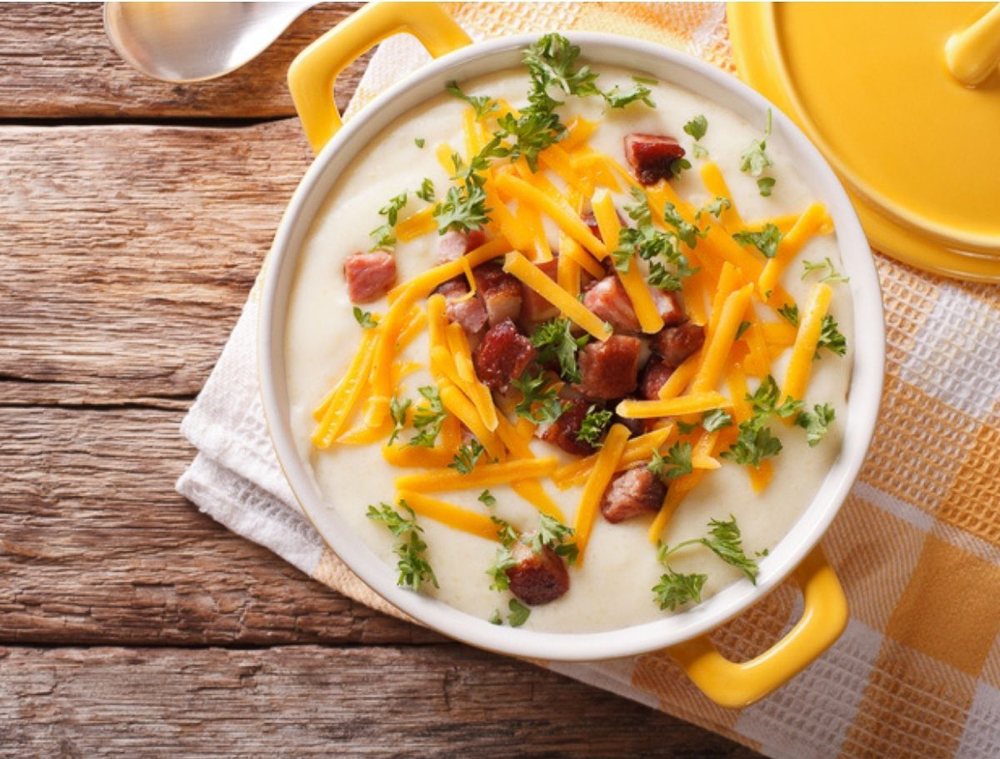 A white bowl of loaded potato soup with cheese, bacon and herbs on top of it with a yellow lid to the side.