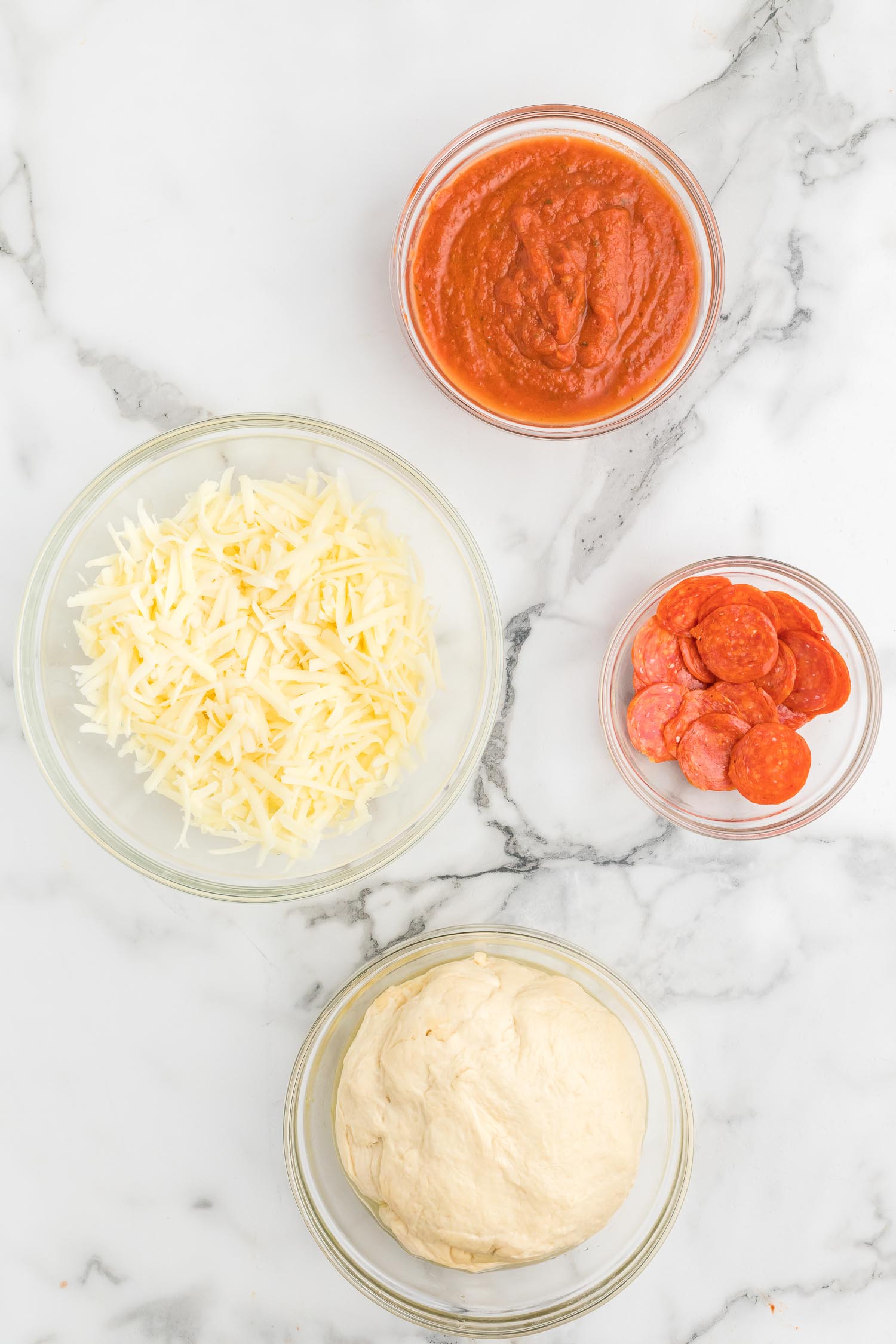 Glass bowls with pizza toppings: pepperoni, shredded cheese, tomato sauce, homemade pizza dough.
