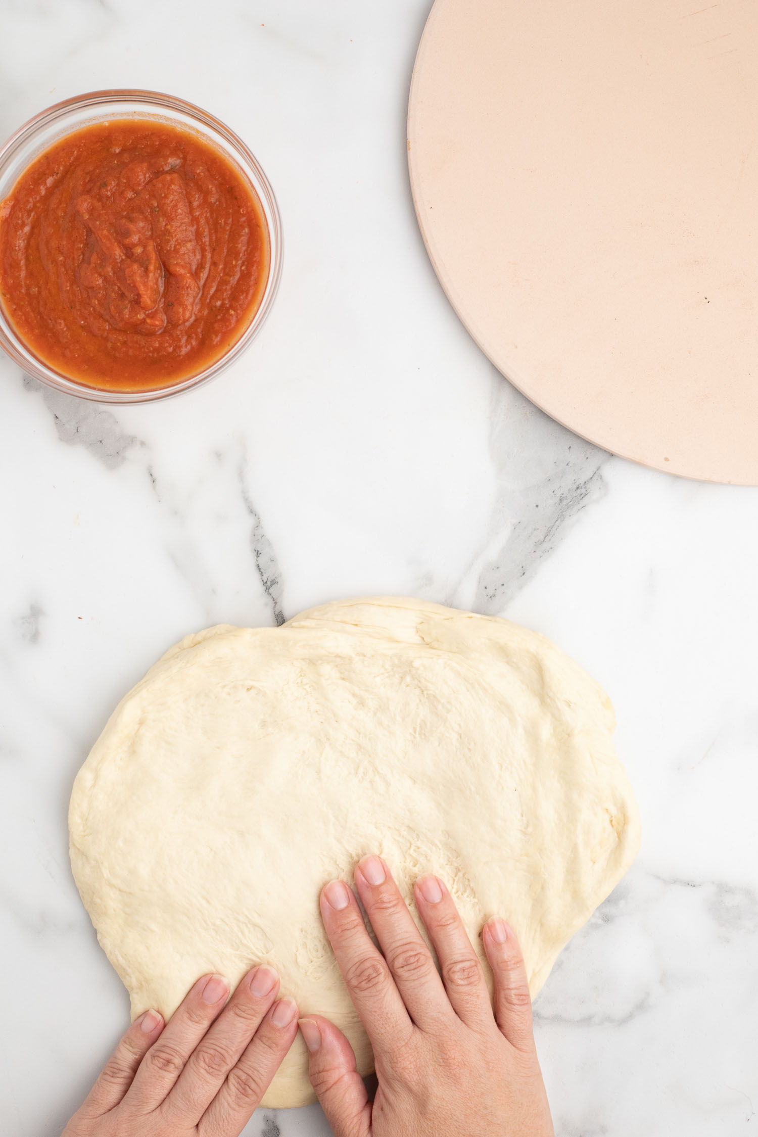 A pizza stone with a glass bowl of tomato sauce with a bunch of homemade pizza dough with two hands rolling it out.