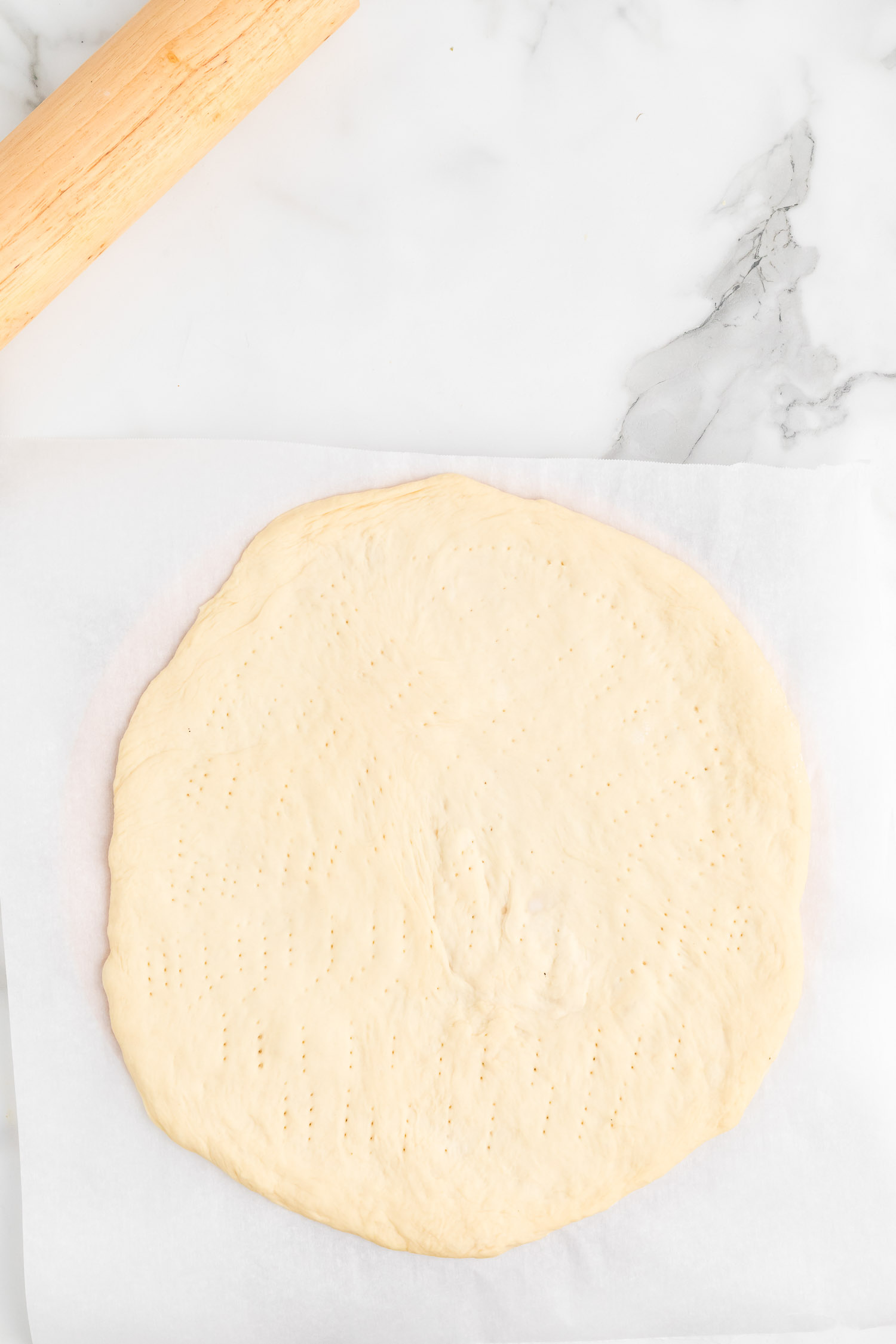 A piece of parchment paper with a rolled out pizza dough poked all over with a wooden rolling pin.