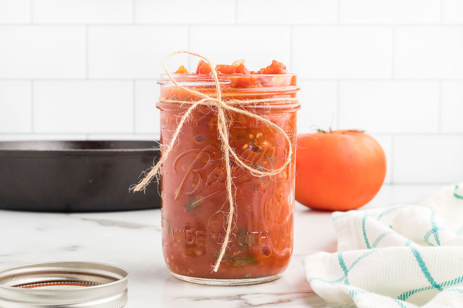 A glass jar with a bow on it filled with homemade tomato sauce  with a black pan and whole tomato behind it.
