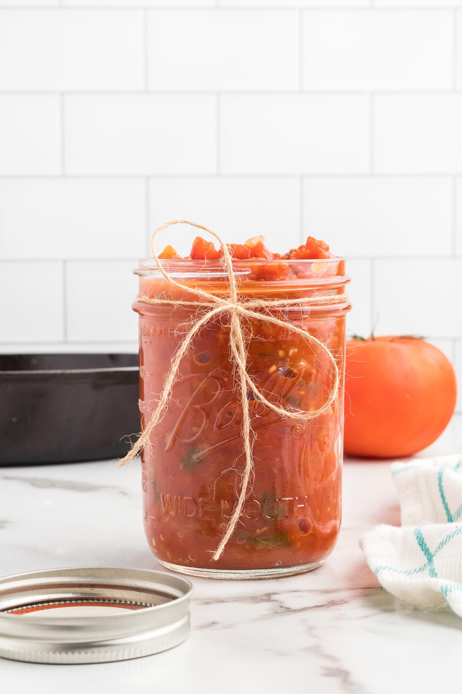 A glass jar with a bow at the front filled with homemade tomato sauce with a tomato, silver lid, and black bowl in the background.