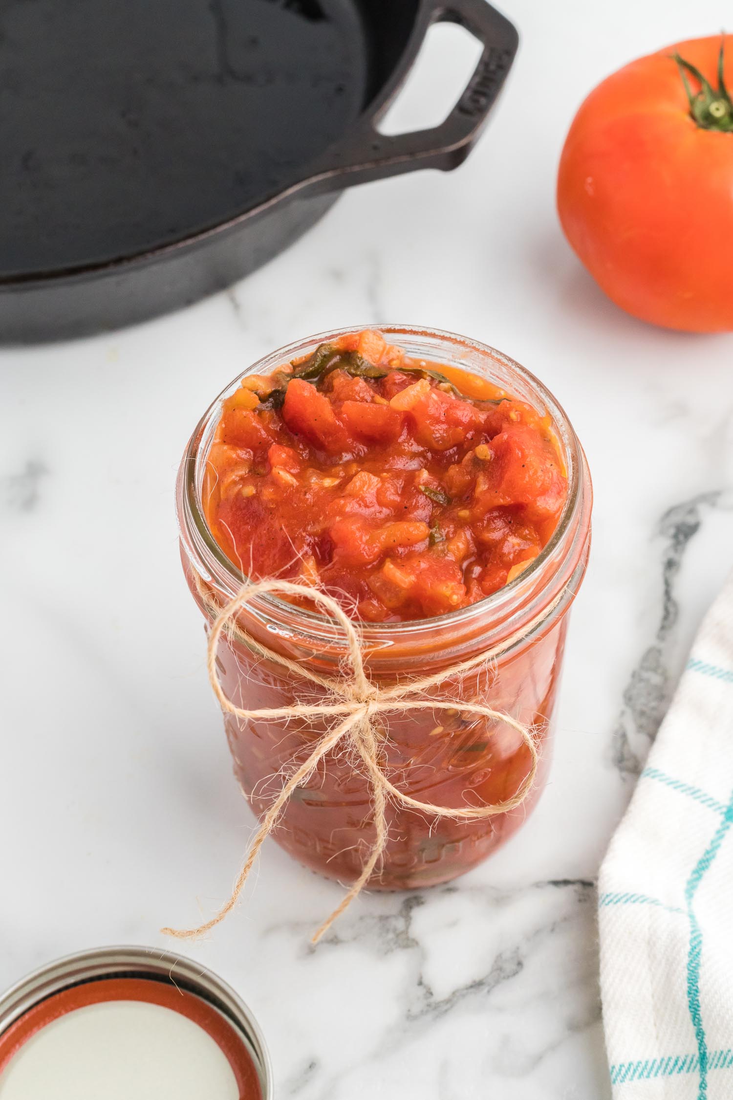 A glass jar with tomato sauce in it with a brown bow tied around the jar with the silver lid and a green and white towel next to it.