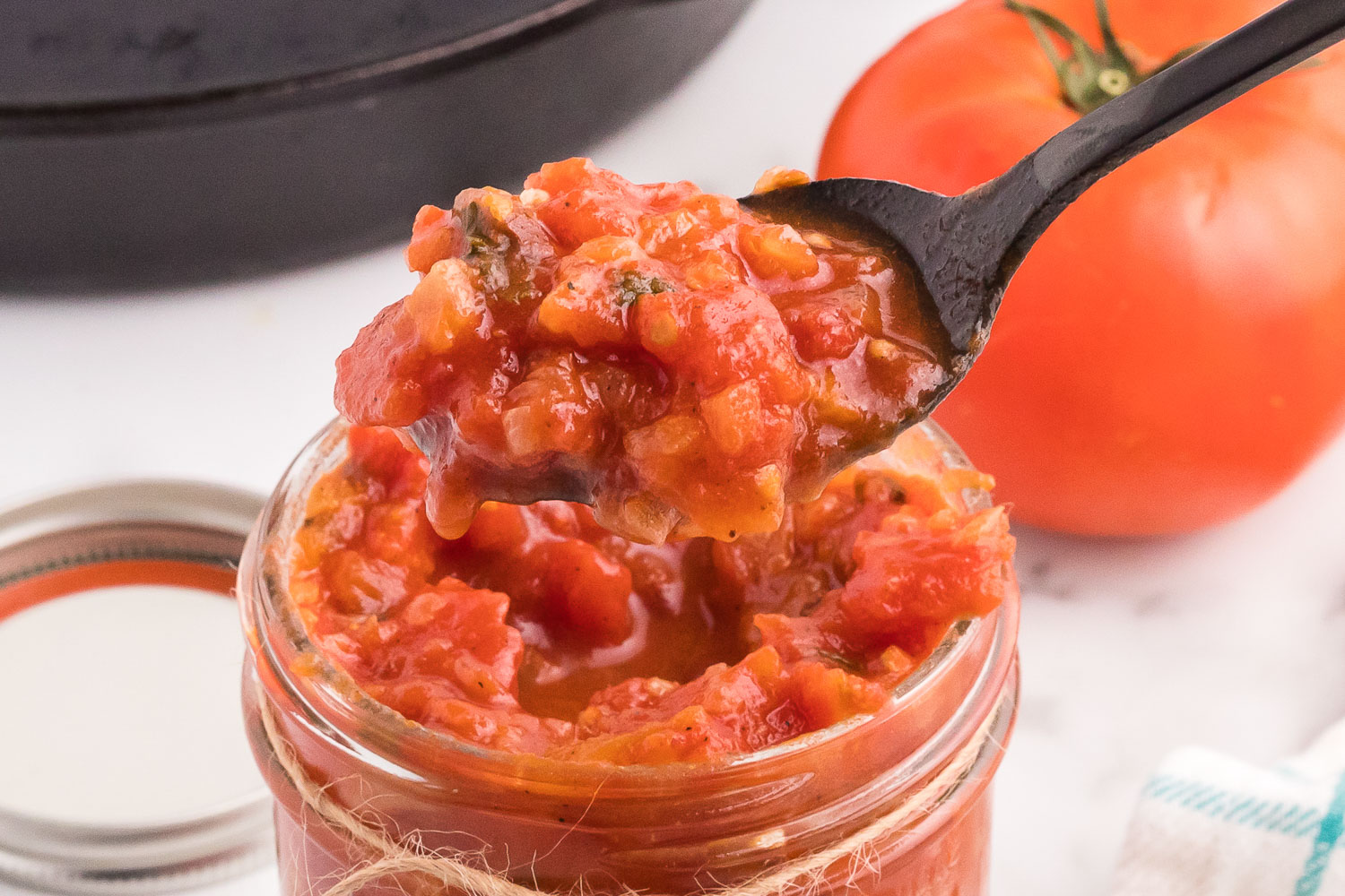a glass jar filled with tomato sauce with a black spoon lifting some of it up with a tomato and a silver lid in the background.