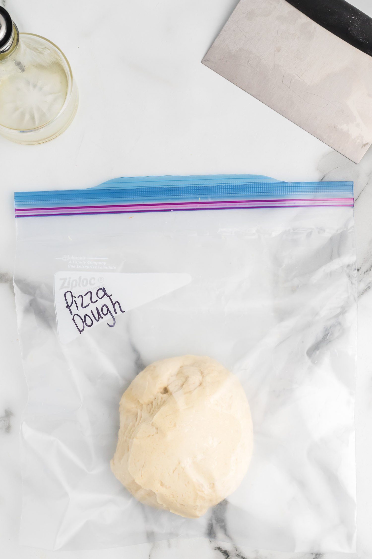 A resealable bag with a ball of dough with the words pizza dough on it, There is oil and a pizza cutter to the side. 