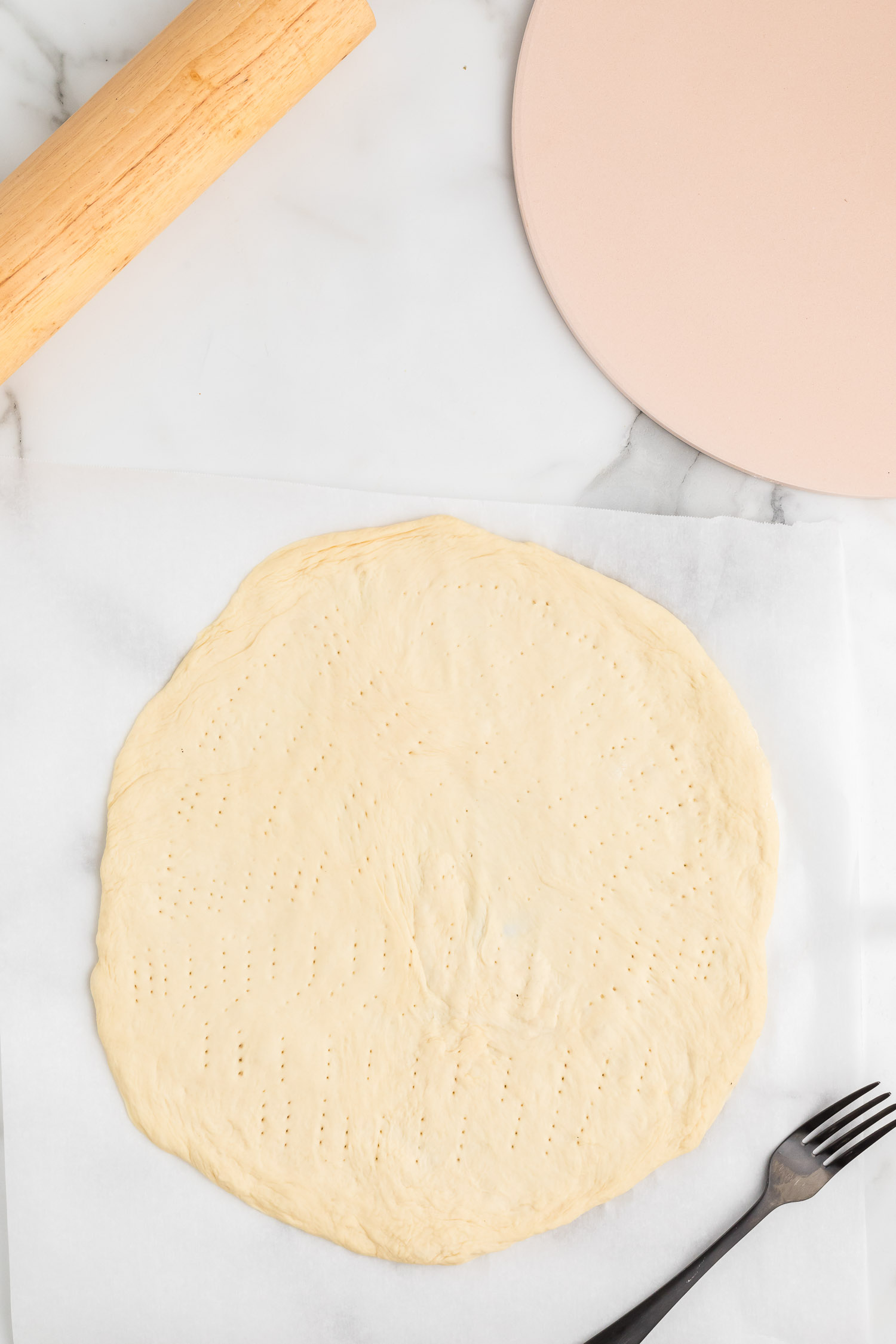 A pizza dough rolled out on parchment paper poked with fork tines with a pizza stone and a black fork on the side.