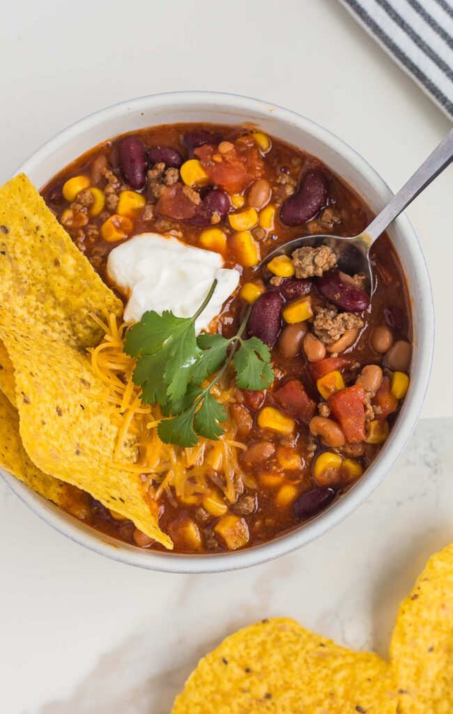 beautiful photo of easy Taco Soup recipe in a white bowl with a spoon, topped with tortilla chips, sour cream and cilantro